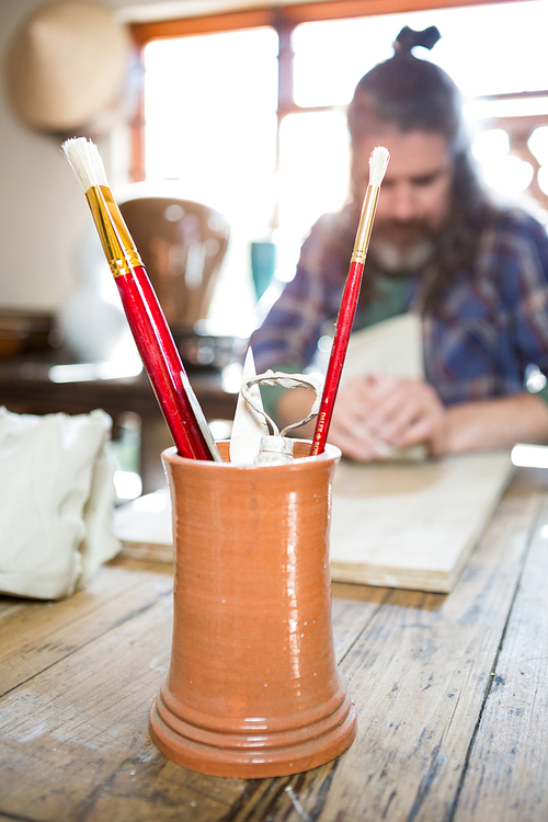 Close-up of paintbrush and work tool in pot with potter working in background