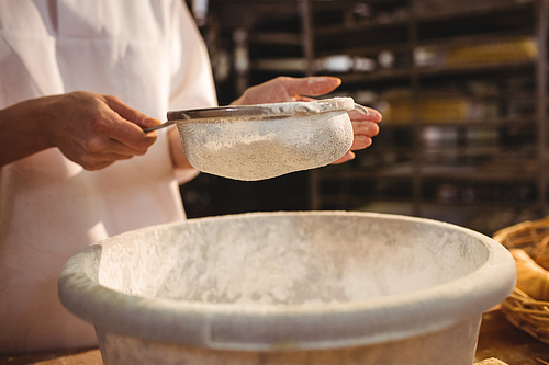 Mid-section of female baker sifting flour through a sieve in bakery shop