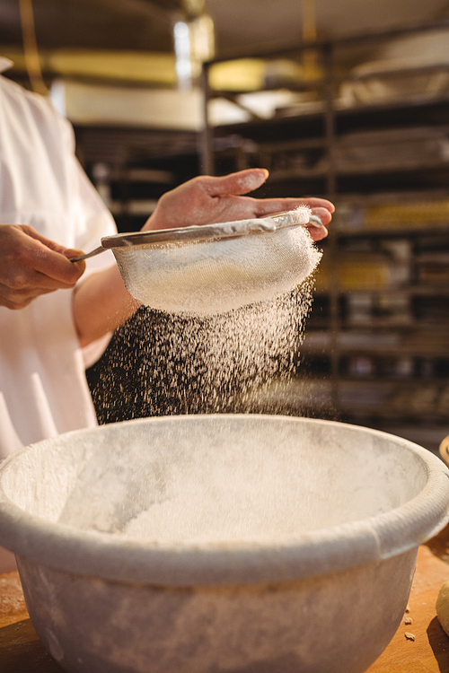 Mid-section of female baker sifting flour through a sieve in bakery shop