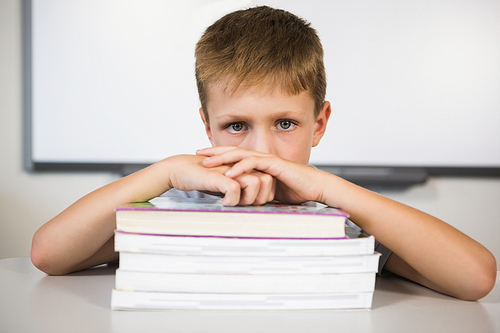 Portrait of sad schoolboy leaning on stack of books in class room at school