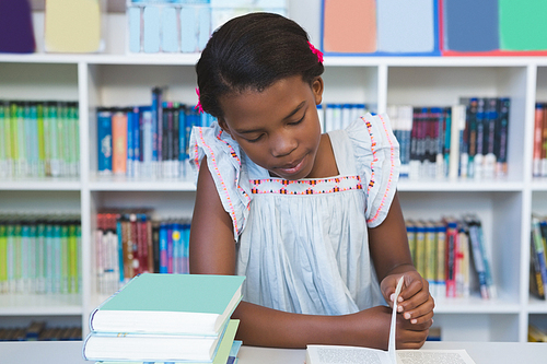Schoolgirl sitting on table and reading book in library at school