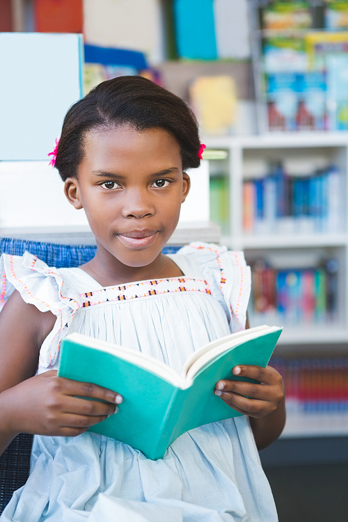 Portrait of schoolgirl sitting on chair and reading book in library at school