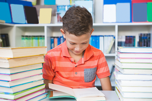 Schoolboy sitting on table and reading book in library at school