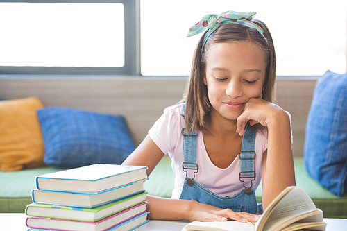 Schoolgirl sitting on table and reading book in library at school