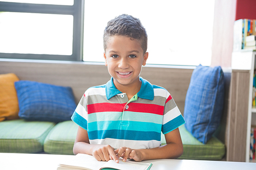 Portrait of schoolboy sitting on table and reading book in library at school