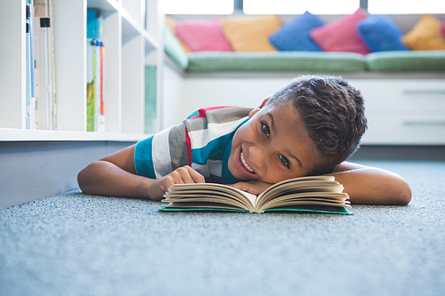 Portrait of schoolboy lying on floor and reading a book in library at school
