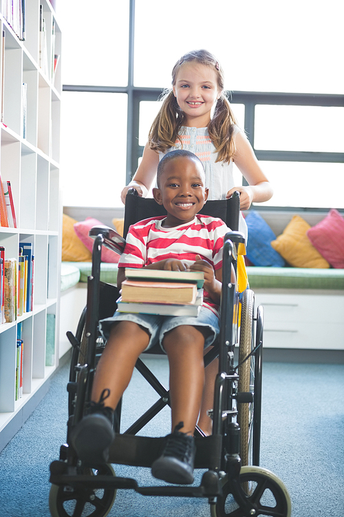 Portrait of happy schoolgirl carrying schoolboy in wheelchair in library