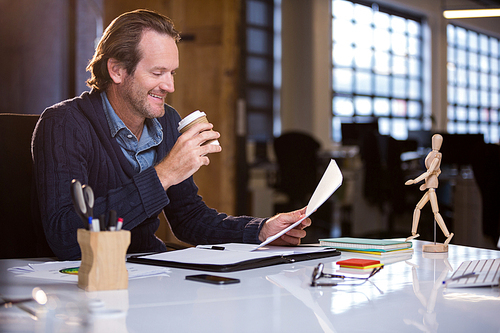 Happy businessman having coffee while reading document at desk in creative office