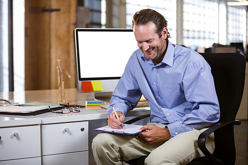 Businessman smiling while writing on clipboard by desk in creative office