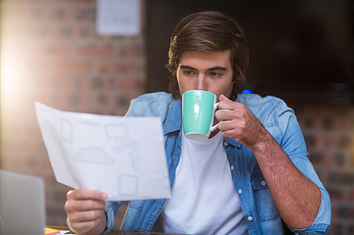 Young businessman having coffee while reading document in creative office