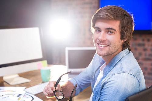 Portrait of young businessman holding eyeglasses at desk in creative office