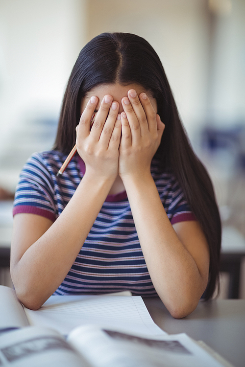 Stressed schoolgirl doing covering her face in classroom at school