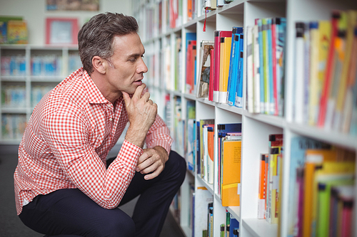 Thoughtful school teacher selecting book in library at school