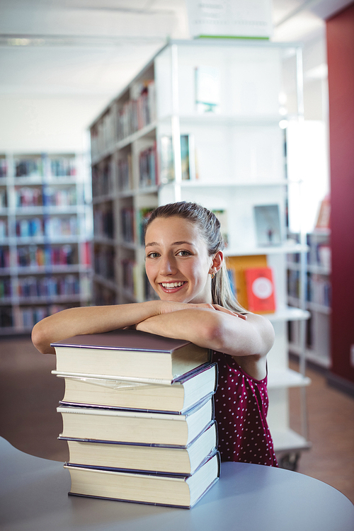 Portrait of happy schoolgirl leaning of stacked books in library at school