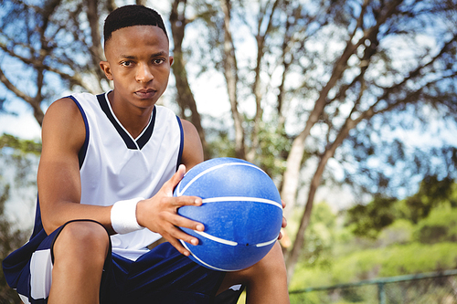 Portrait of male teenager with basketball sitting on bench in court