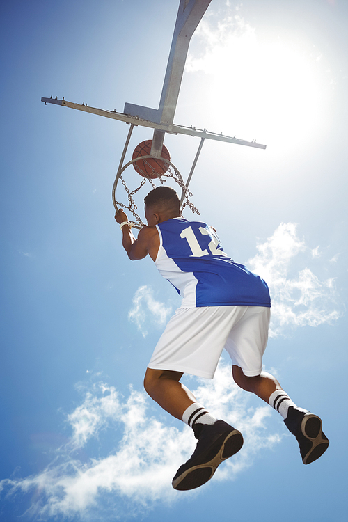Rear view of male teenager hanging on basketball hoop against blue sky
