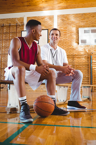 Smiling coach and player talking while sitting on bench in court