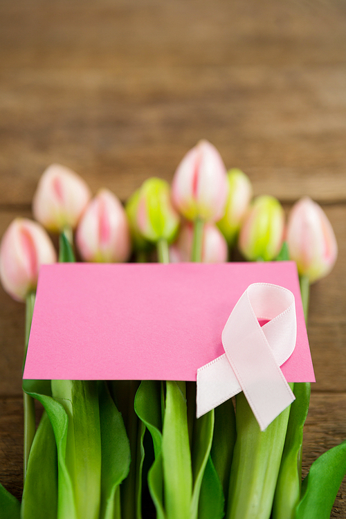 Close-up of pink Breast Cancer Awareness ribbon with blank card on fresh tulips over wooden table