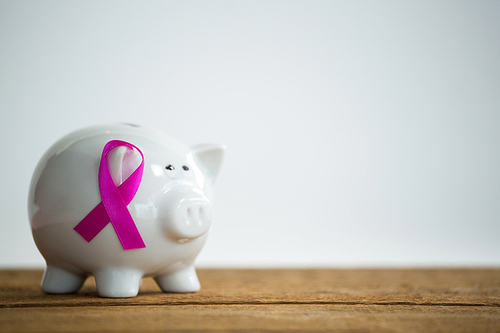 Close-up of pink Breast Cancer Awareness ribbon on piggybank over wooden table against white background