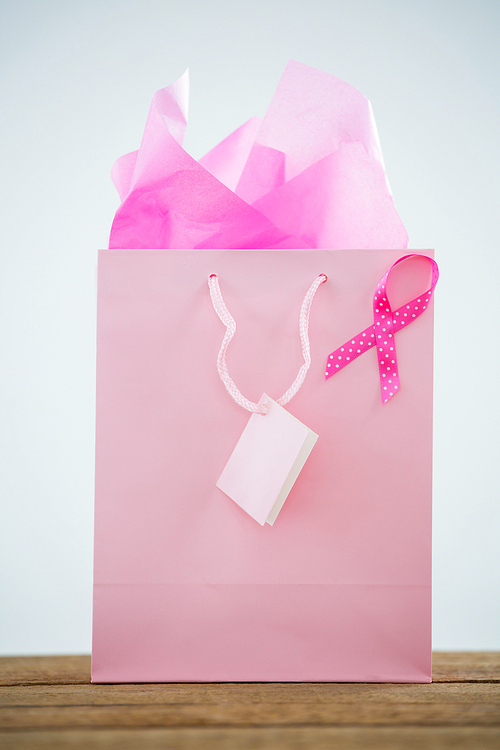 Close-up of pink Breast Cancer Awareness ribbon on shopping bag over wooden table against white background