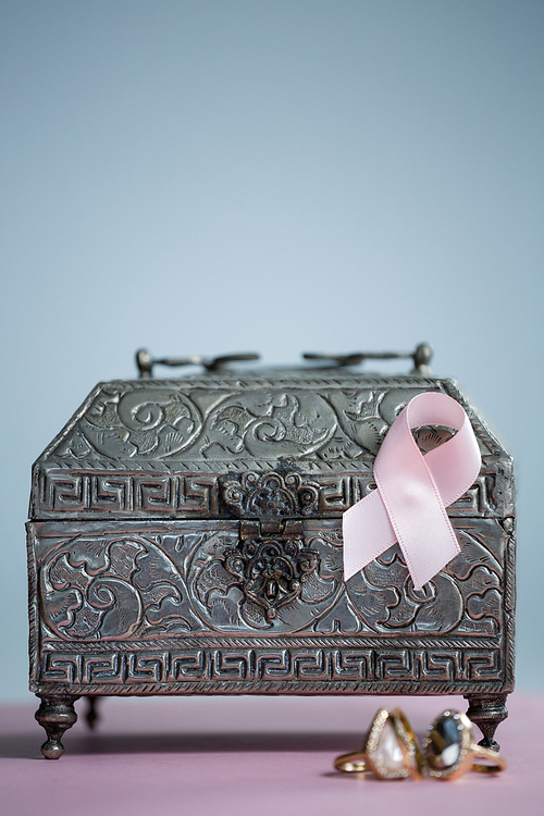 Close-up of pink Breast Cancer Awareness ribbon with rings and chest on table against white background