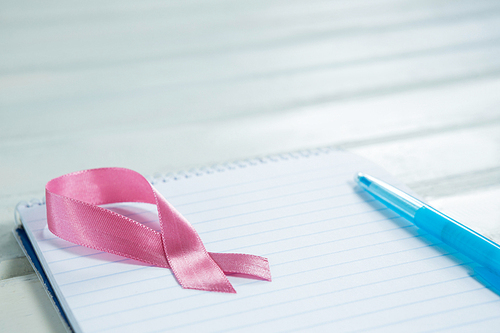 High angle view of pink Breast Cancer Awareness ribbon and spiral notepad with pen on wooden table