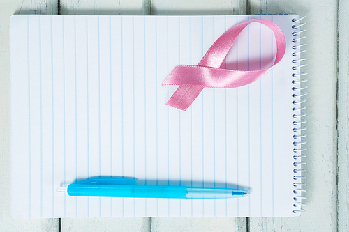 Overhead view of pink Breast Cancer Awareness ribbon and spiral notepad with pen on wooden table