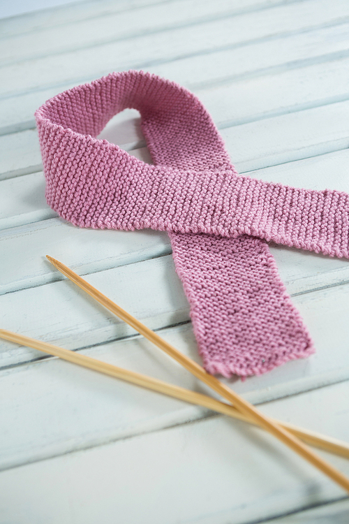 Close-up of pink woolen Breast Cancer Awareness ribbon by crochet needles on white wooden table