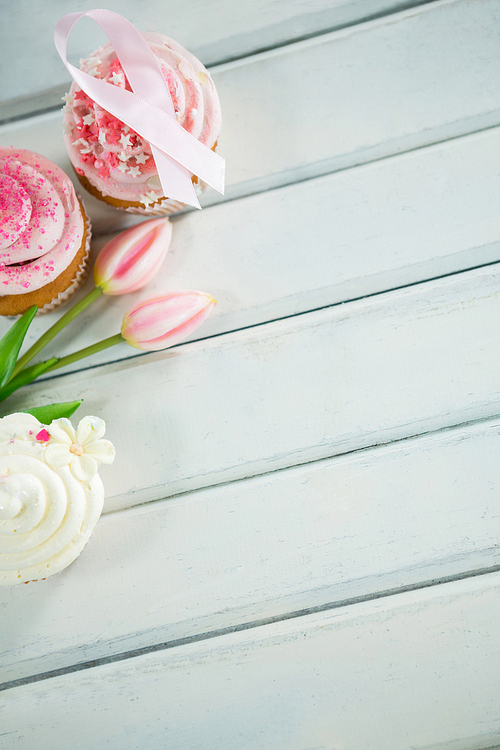 Overhead view of Breast Cancer Awareness pink ribbons on cupcakes with tulips over white wooden table