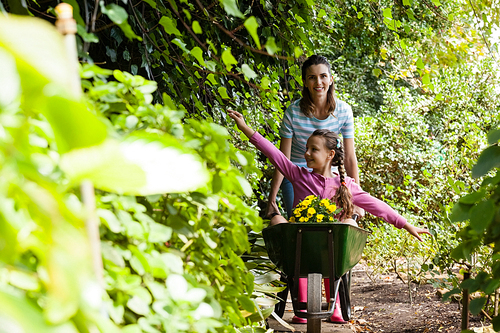 Smiling woman pushing girl sitting with outstretched in wheelbarrow on footpath amidst plants at garden