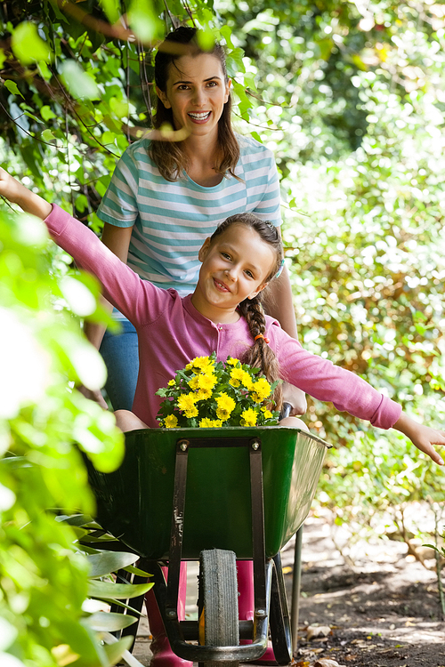 Girl enjoying with arms outstretched while smiling mother pushing wheelbarrow on footpath by plants