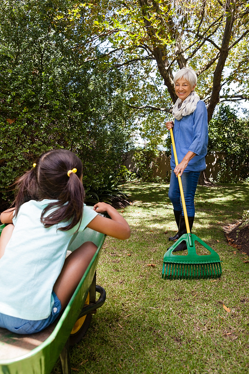 Grandmother holding rake while looking granddaughter sitting in wheelbarrow at backyard
