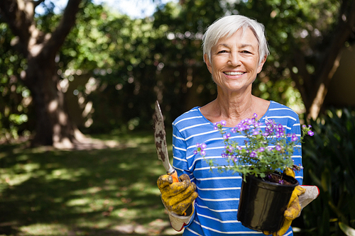 Portrait of senior woman holding potted plant and trowel at backyard