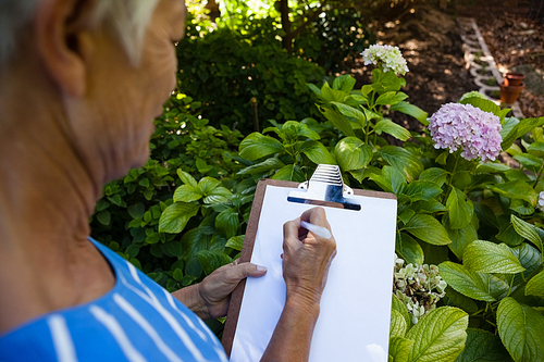 Midsection of senior woman writing on clipboard against plants at backyard
