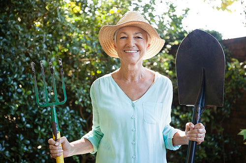 Portrait of smiling senior woman holding garden fork and shovel at backyard
