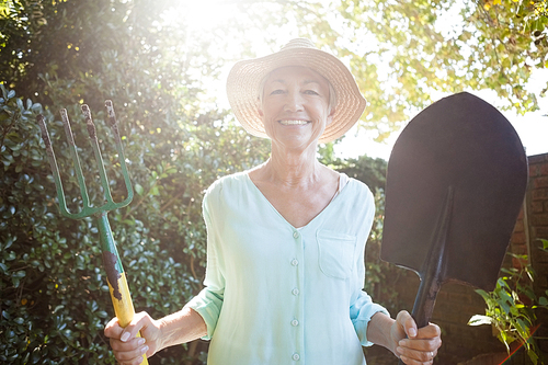 Back lit portrait of smiling senior woman holding garden fork and shovel at backyard