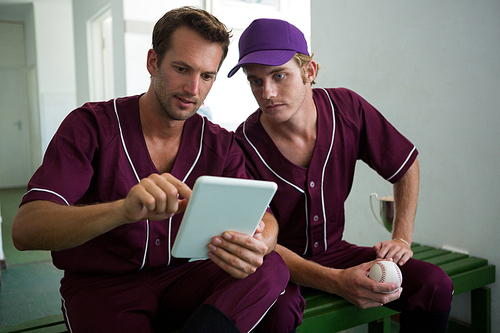 Baseball players using digital tablet while sitting on bench at locker room