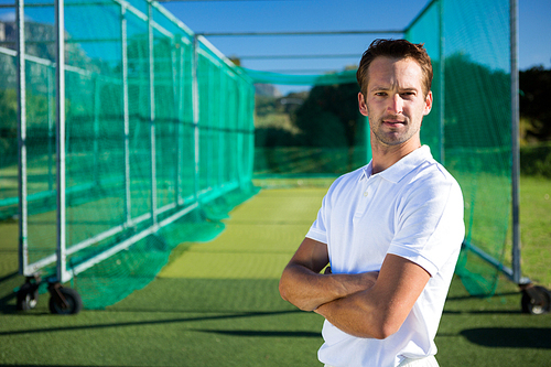 Portrait of young cricketer with arms crossed standing against net on field