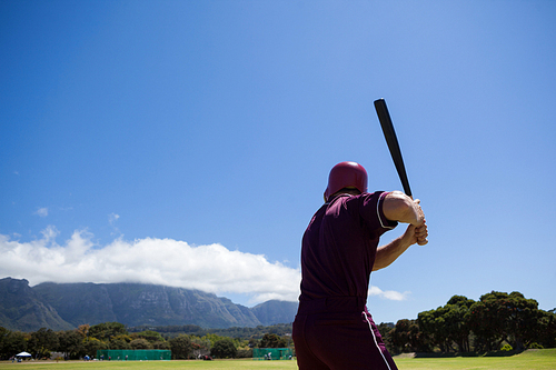 Rear view of baseball player with bat standing at field against blue sky