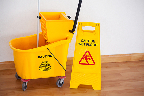 Yellow sign board with mop bucket on wooden floor against wall