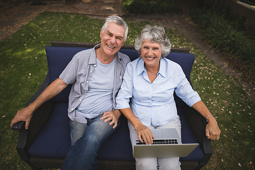 High angle portrait of smiling senior couple sitting together with laptop on couch in backyard