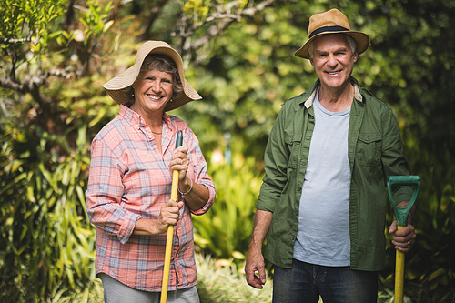 Portrait of smiling senior couple holding tools while standing at yard