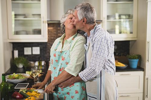Loving senior couple kissing while cooking in kitchen at home