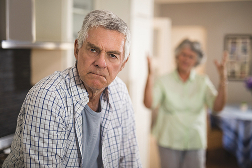 Portrait of unsmiling senior man with angry woman in background at home