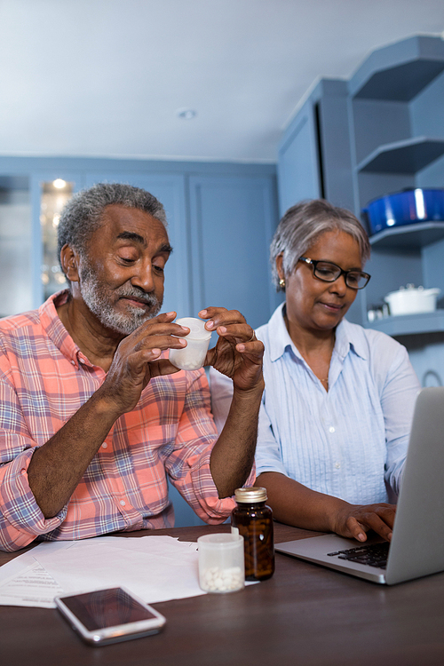 Man looking at medicine while sitting by woman using laptop computer in kitchen at home