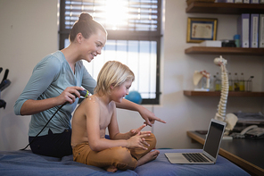 Smiling female therapist pointing at laptop while scanning shoulder of shirtless boy in hospital ward