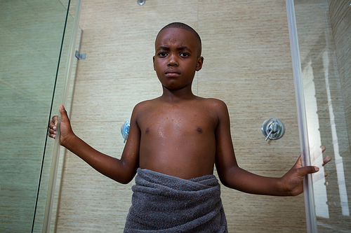 Low angle portrait of shirtless boy wrapped in towel standing amidst glass against wall at bathroom