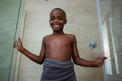 Low angle portrait of smiling shirtless boy wrapped in gray towel standing amidst glass against wall at bathroom