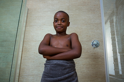 Low angle portrait of shirtless boy wrapped in towel standing with arms crossed amidst glass against wall at bathroom