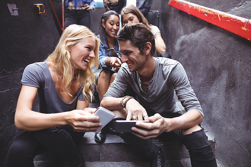 Cheerful friends using mobile phones while sitting on steps at nightclub